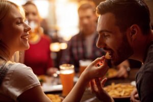 Young couple enjoying in lunch with their friends in a restaurant. Young man is being fed by his girlfriend with nacho chips.
